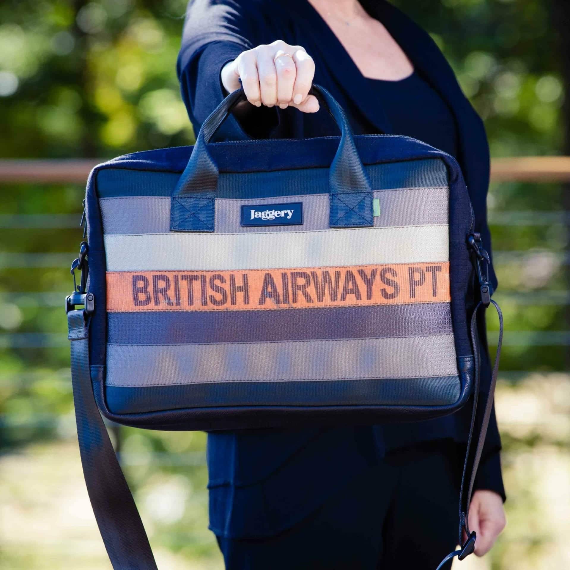 Upcycled Executive Urban Briefcase a briefcase held out in a woman's hand made from upcycled cargo belts, with the middle one saying "British Airways"