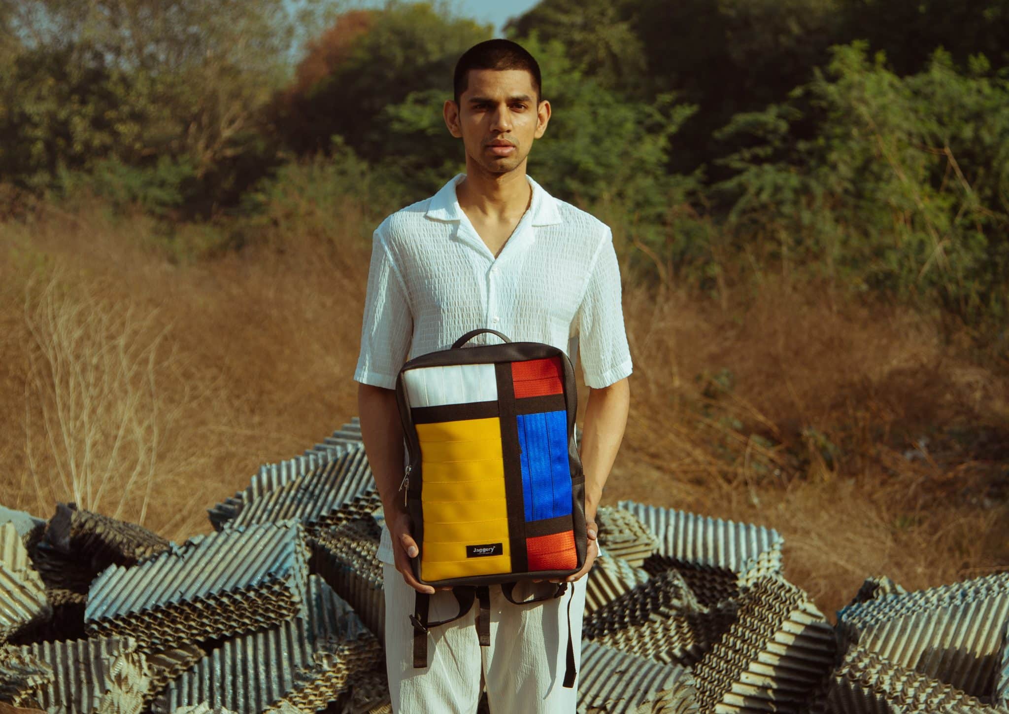Jaggery Upcycled Seat Belt Backpack a man stands holding a colorful backpack made from upcycled seat belts