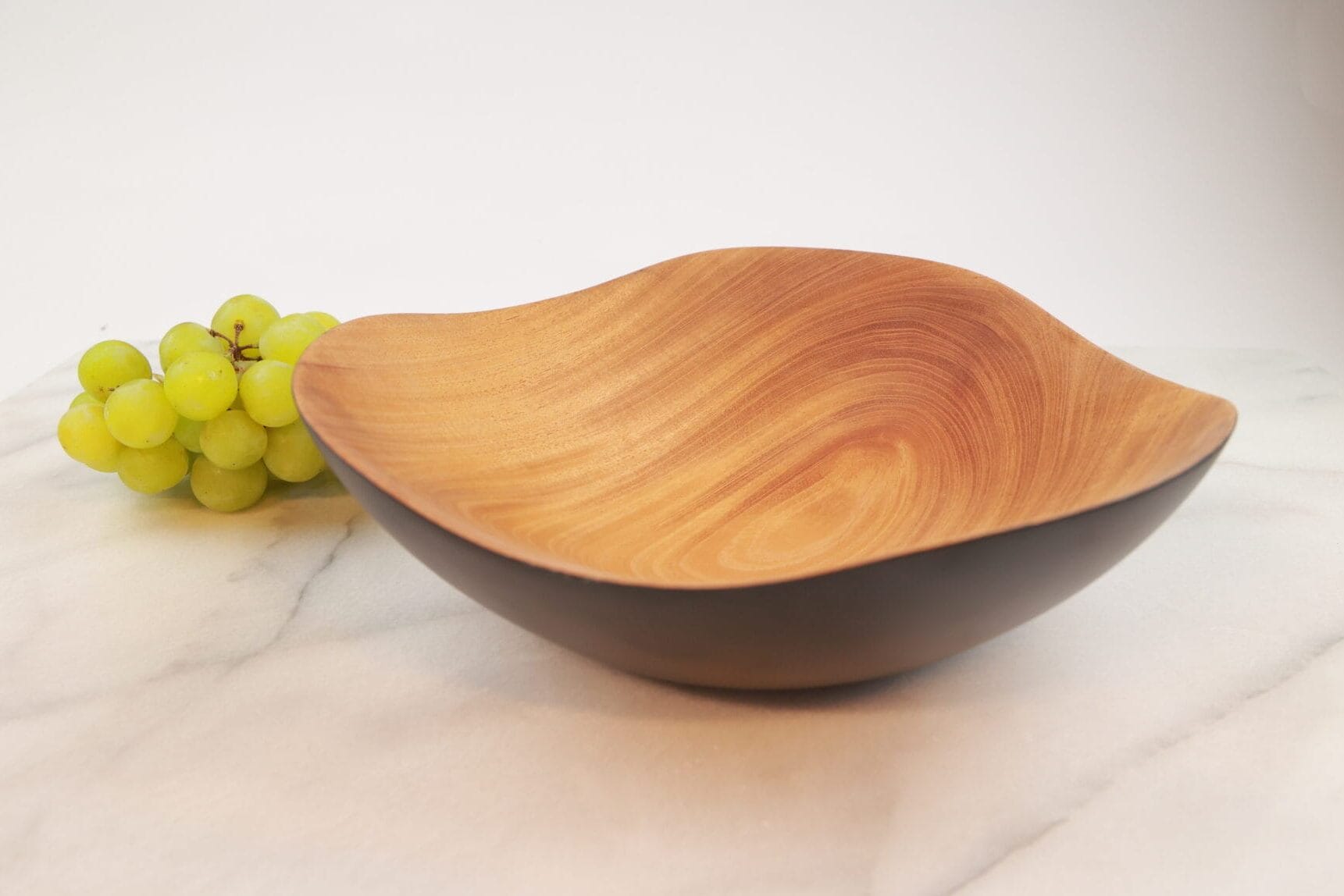 Itza Wood charred Wave Bowl next to grapes wave shaped wooden bowl with charred outer edge seated next to a bundle of green grapes